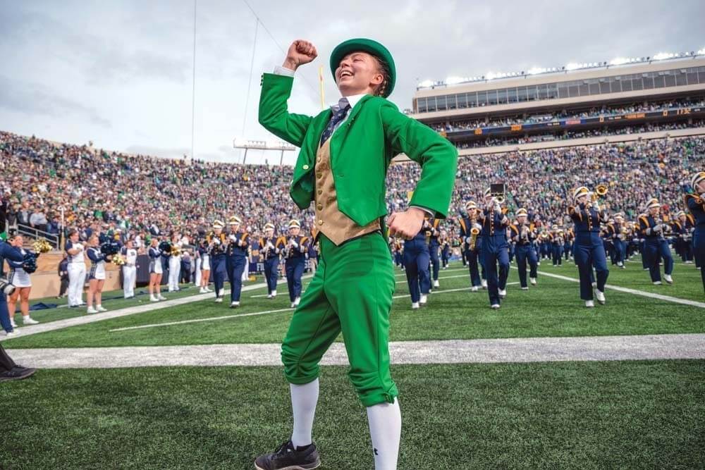 Kylee in the green Leprechaun suit, smiling with a fist raised, on the field at Notre Dame Stadium with the marching band behind her.